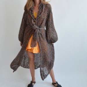 May include: A long, brown, knit cardigan with a tie belt. The cardigan has a loose fit and a slightly flared hem. The cardigan is made of a soft, lightweight yarn and has a subtle shimmer.