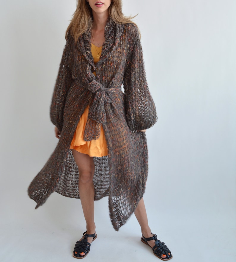 May include: A long, brown, knit cardigan with a tie belt. The cardigan has a loose fit and a slightly flared hem. The cardigan is made of a soft, lightweight yarn and has a subtle shimmer.