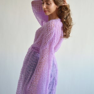 May include: A light purple knitted cardigan with a loose fit and long sleeves. The cardigan has a delicate openwork pattern and a tie belt at the waist.