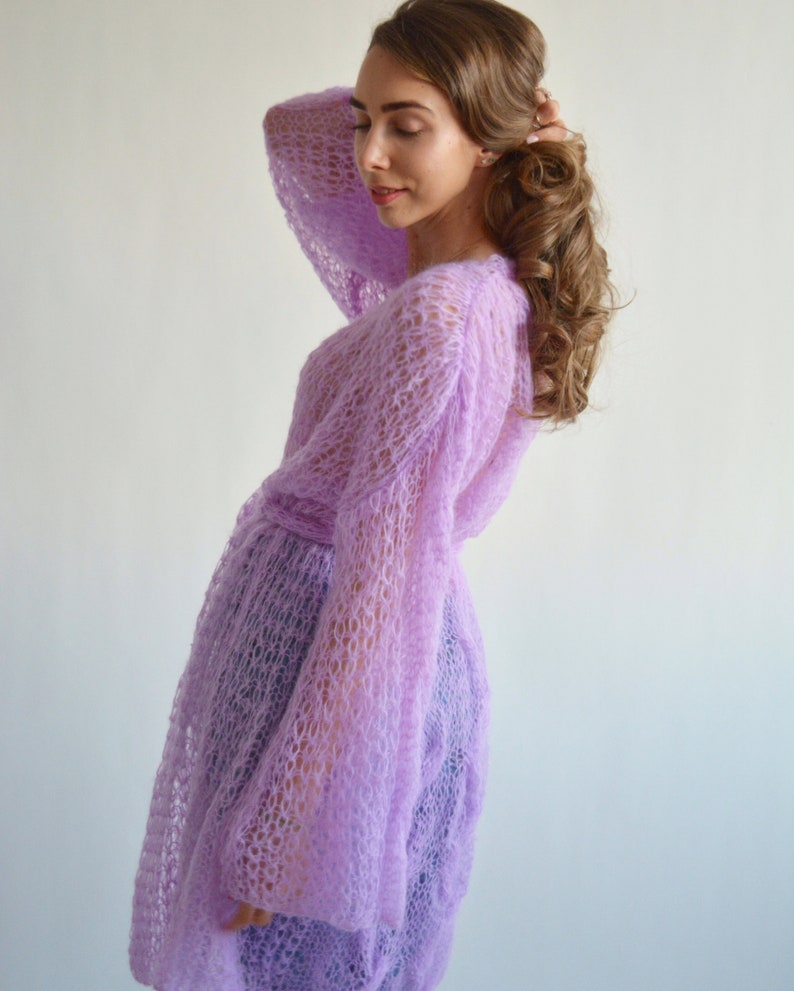 May include: A light purple knitted cardigan with a loose fit and long sleeves. The cardigan has a delicate openwork pattern and a tie belt at the waist.