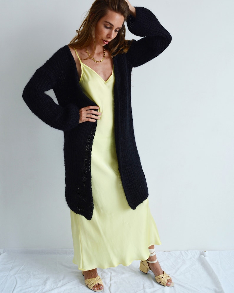 May include: A black knitted cardigan with long sleeves and a loose fit, worn over a pale yellow slip dress. The model is wearing the cardigan open, with the sleeves rolled up.