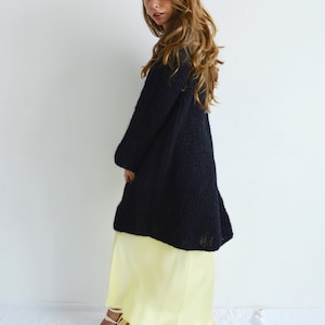 Op de afbeelding: Een zwarte, lange gebreide cardigan met een losse pasvorm en een gele satijnen maxi-rok. Het model draagt de cardigan open en de rok valt soepel.