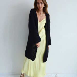 Op de afbeelding: Een vrouw draagt een zwarte gebreide cardigan over een lichtgele slipdress. Ze draagt witte sandalen met een geweven band.