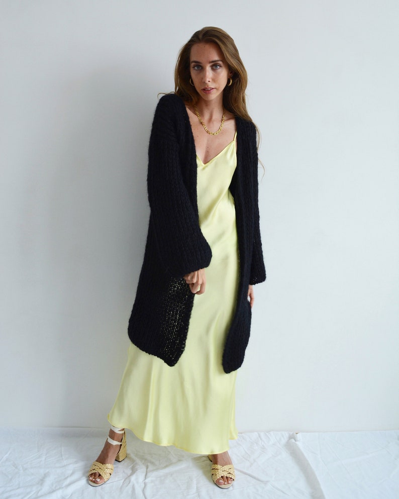 Op de afbeelding: Een vrouw draagt een zwarte gebreide cardigan over een lichtgele slipdress. Ze draagt witte sandalen met een geweven band.