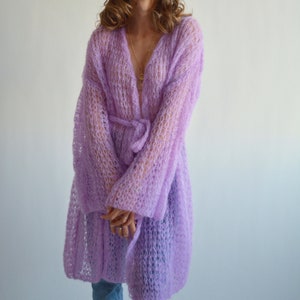 May include: A long, light purple knitted cardigan with a tie belt and long sleeves. The cardigan is made of a loose, open knit fabric.
