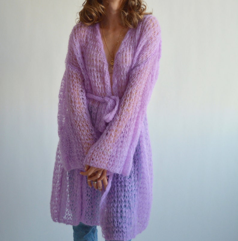 May include: A long, light purple knitted cardigan with a tie belt and long sleeves. The cardigan is made of a loose, open knit fabric.