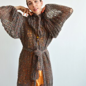 May include: A long, brown, knitted cardigan with a loose fit and a tie belt. The cardigan has a textured, open-weave design and is made of a soft, fuzzy yarn.