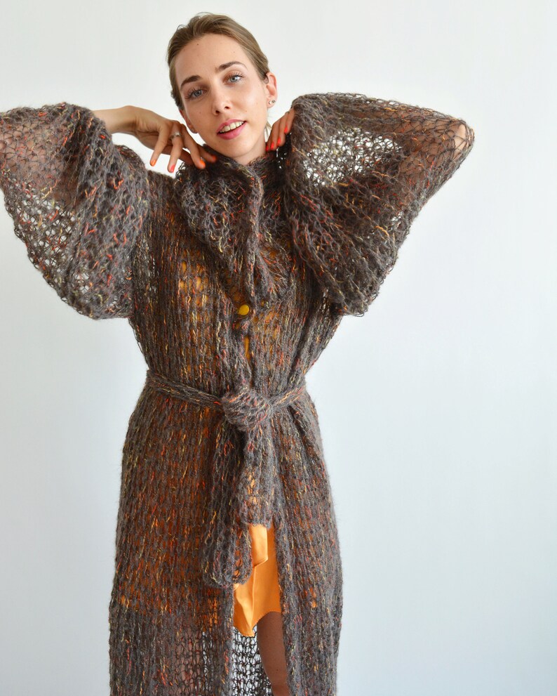 May include: A long, brown, knitted cardigan with a loose fit and a tie belt. The cardigan has a textured, open-weave design and is made of a soft, fuzzy yarn.