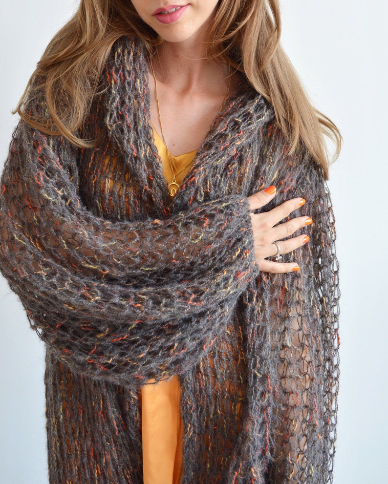 May include: A brown, orange, and gray knitted cardigan with a loose, open weave. The cardigan is worn open and has a long, flowing style.