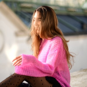 以下が含まれることがあります： ピンクのふわふわしたセーターと白のドット柄の黒いタイツを履いた女性がコンクリートの表面に座っています。彼女は長い茶色の髪をしていて、下を見ています。