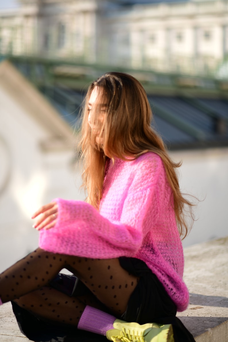 以下が含まれることがあります： ピンクのふわふわしたセーターと白のドット柄の黒いタイツを履いた女性がコンクリートの表面に座っています。彼女は長い茶色の髪をしていて、下を見ています。