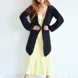 Op de afbeelding: Een zwarte, lange cardigan met een losse pasvorm en een V-hals. De cardigan wordt gedragen over een gele satijnen slipdress.