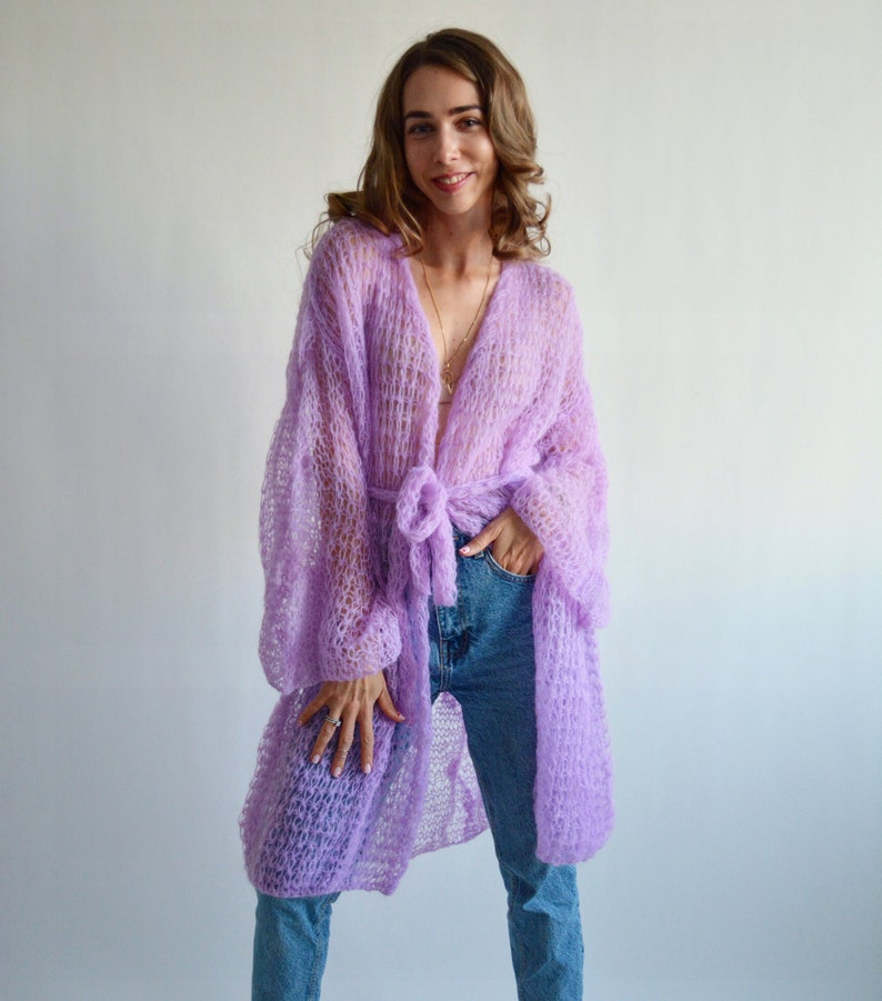 May include: A long, light purple knitted cardigan with a tie belt. The cardigan is made of a loose, open weave and has a relaxed fit.
