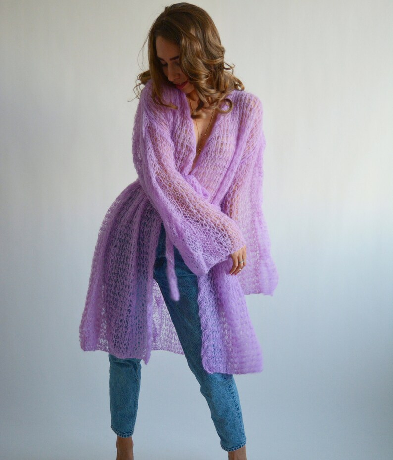 May include: A long, light purple knitted cardigan with a loose fit and a tie belt. The cardigan is made of a delicate, open-weave fabric.