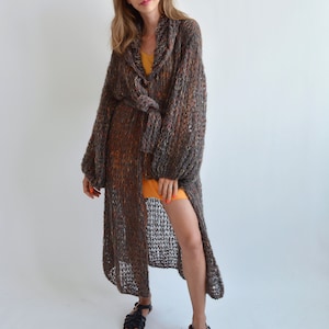 May include: A long, brown, knit cardigan with a tie belt. The cardigan has a loose fit and a slightly flared hem. The model is wearing the cardigan over a yellow dress and black sandals.
