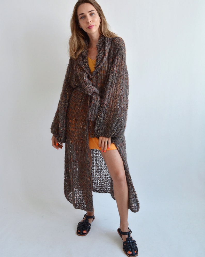 May include: A long, brown, knit cardigan with a tie belt. The cardigan has a loose fit and a slightly flared hem. The model is wearing the cardigan over a yellow dress and black sandals.