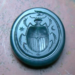 Puede incluir: Talla de piedra ovalada verde oscuro con un diseño detallado de escarabajo. El escarabajo está rodeado por un borde circular con una estrella, una luna creciente y otros elementos decorativos. La talla tiene un acabado pulido.
