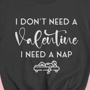 Op de afbeelding: Zwart sweatshirt met witte tekst die "Ik heb geen Valentijn nodig, ik heb een dutje nodig" zegt en een kleine illustratie van een persoon die op een bed slaapt.