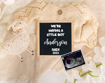 May include: A white knitted baby cardigan, a white crocheted lamb toy, a white orchid, and a black letter board with the text "We're having a little boy Anderson March 2023" and a black and white ultrasound image.
