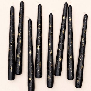 May include: Eight black taper candles arranged vertically, each featuring a metallic gold celestial pattern of stars, crescent moons, and small dots. The candles have a smooth, cylindrical shape with a pointed top and a flat base.