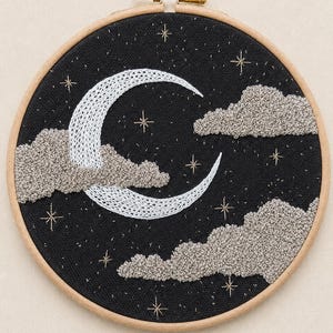 Op de afbeelding: Een ronde houten borduurring met een zwarte stoffen achtergrond. Het ontwerp toont een witte halve maan, gestructureerde lichtgrijze wolken en kleine goudkleurige geborduurde sterren verspreid over de nachtelijke hemel.