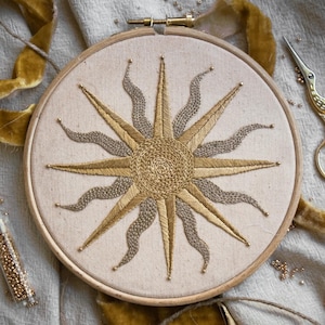 Peut inclure: Un tambour à broder circulaire en bois contenant un panneau en tissu beige avec un motif de soleil cousu à la main. Le soleil présente un motif circulaire central avec des rayons dorés droits et des rayons argentés ondulés alternés.