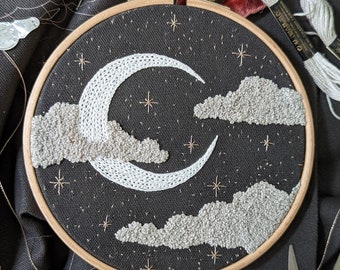 Op de afbeelding: Een ronde houten borduurring met een zwart stoffen paneel voorzien van een handgeborduurd ontwerp van een witte maansikkel, gestructureerde lichtgrijze wolken en kleine goudkleurige stermotieven op een donkere nachtelijke hemel.