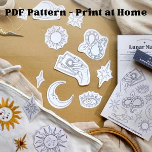 Op de afbeelding: Een verzameling borduurbenodigdheden en patronen. Inclusief een witte canvas tas met een zonontwerp, papieren uitsparingen van hemelse symbolen, een schaar en een boekje met de tekst "Lunar Magic" met "Embroidery Transfers". De tekst "PDF Pattern - Print at Home" is zichtbaar.