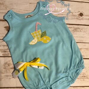 Könnte beinhalten: Ein hellblauer Baby-Strampler mit einem gelben Limonade-Applikationen. Der Strampler hat eine gelbe Schleife mit weißen Punkten, die an der Seite befestigt ist.