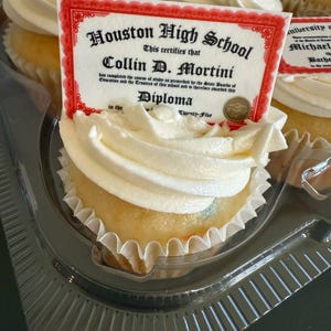 Może przedstawiać: Babeczki z białym lukrem i dekoracją w kształcie dyplomu. Na dyplomie widnieje napis "Houston High School" i "Collin D. Mortini". Babeczki znajdują się w przezroczystym plastikowym pojemniku.