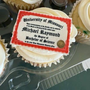 Może przedstawiać: Babeczka z białym lukrem i dekoracyjnym, jadalnym dyplomem. Na dyplomie widnieje napis "University of Missouri" i "Michael Raymond, Bachelor of Science, in the year Two Thousand Twenty-Five". Babeczka znajduje się w przezroczystym plastikowym pojemniku.