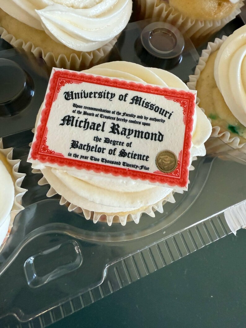 Może przedstawiać: Babeczka z białym lukrem i dekoracyjnym, jadalnym dyplomem. Na dyplomie widnieje napis "University of Missouri" i "Michael Raymond, Bachelor of Science, in the year Two Thousand Twenty-Five". Babeczka znajduje się w przezroczystym plastikowym pojemniku.