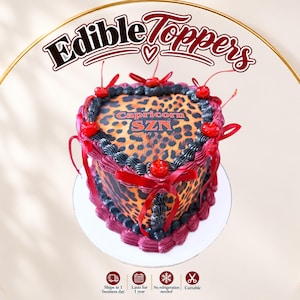 May include: A heart-shaped cake topper featuring a brown and black leopard print pattern with the text Capricorn SZN in orange. The topper is decorated with dark red and black frosting borders, red cherries, and red ribbon bows.