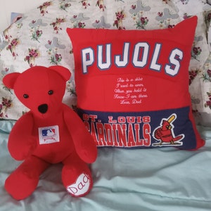 May include: Red teddy bear with a baseball logo on its chest, sitting on a white and floral bedspread next to a red and blue pillow with the words "PUJOLS" and "ST. LOUIS CARDINALS" on it. The pillow has a small red cardinal bird graphic on it. The pillow has a message that reads "This is a shirt I used to wear. When you hold it Know I am there Love, Dad."
