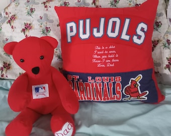 May include: Red teddy bear with a baseball logo on its chest, sitting on a white and floral bedspread next to a red and blue pillow with the words "PUJOLS" and "ST. LOUIS CARDINALS" on it. The pillow has a small red cardinal bird graphic on it. The pillow has a message that reads "This is a shirt I used to wear. When you hold it Know I am there Love, Dad."