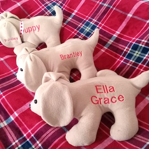 May include: Three plush dog toys on a red and white plaid blanket. The dogs are beige and have the names "Paisley", "Brantley", and "Ella Grace" embroidered on them.