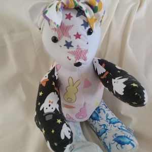 May include: A handmade white teddy bear with a patterned design. The bear has a colorful bow, star and stripe patterns on its face, and arms with a black background and white ghost design. The bear's body features a pink and white bunny pattern and blue crab pattern.