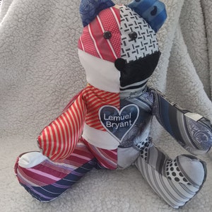 May include: A patchwork teddy bear made from ties, with a heart-shaped tag that reads "Lemuel Bryant". The bear is sitting on a white fluffy surface.