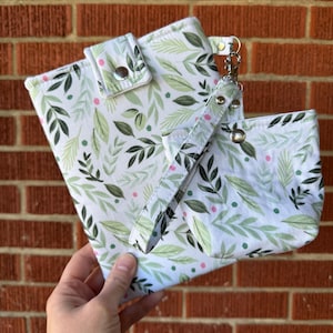 May include: A white fabric pouch and a matching rectangular case featuring a green leaf pattern with small pink dots. Both items have silver metal snap closures, and the pouch includes a detachable wrist strap with a silver metal swivel clasp.