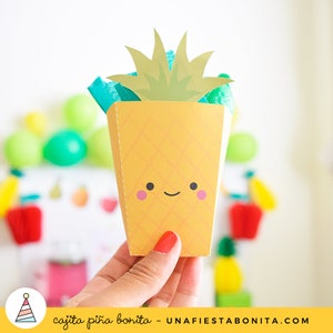 May include: A yellow pineapple-shaped party favor box with a smiling face and pink cheeks. The box is filled with colorful candies and is being held by a hand.