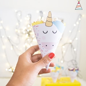 Puede incluir: Un cono blanco con forma de unicornio relleno de un postre amarillo y blanco. El cono tiene un patrón de confeti rosa, azul y blanco. El cono está siendo sostenido por una mano con esmalte de uñas rojo.