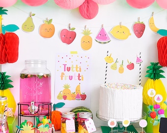 May include: A festive party scene with a fruit theme. Decorations include a banner of smiling fruit cutouts, pink balloons, and paper honeycomb decorations. A cake, cupcakes, and a drink dispenser are on display, along with a sign that says "Tutti Frutti".