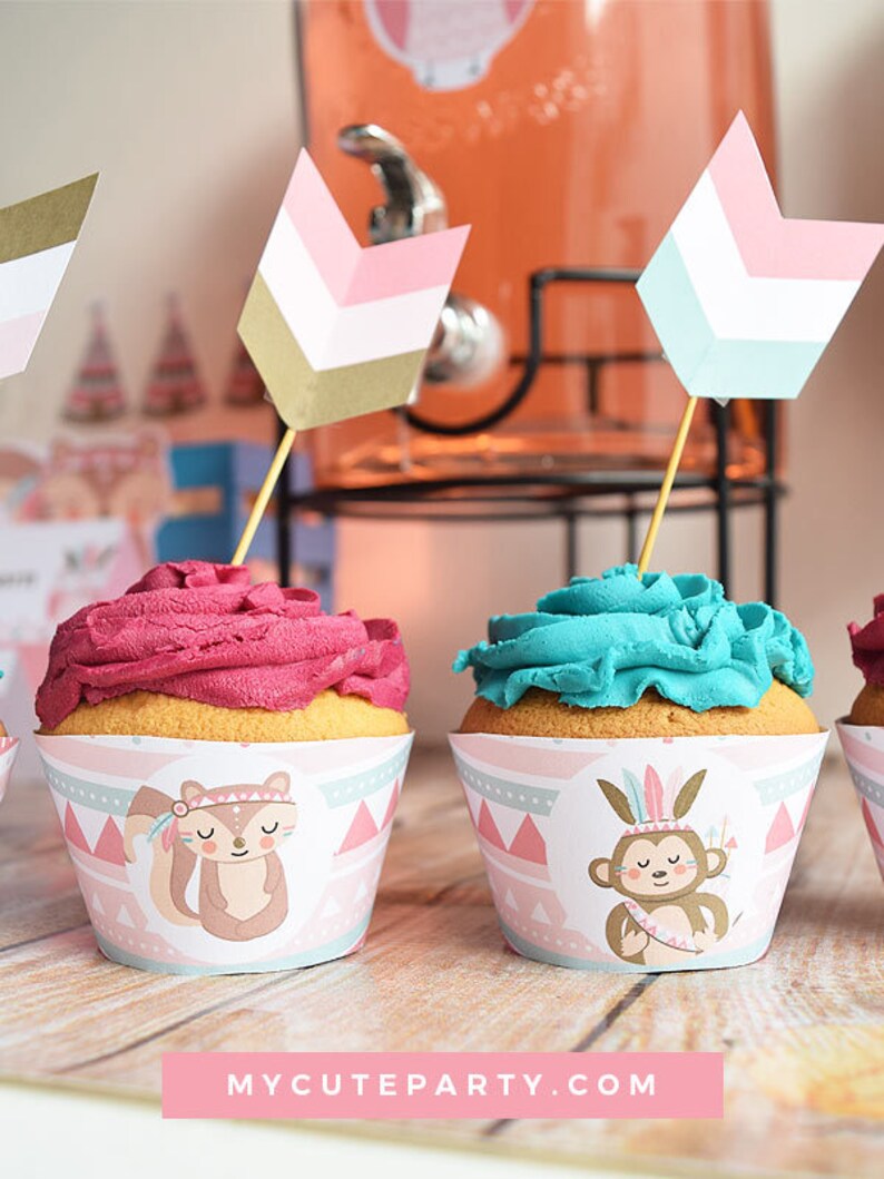 May include: Pink and blue cupcakes with animal-themed wrappers. The wrappers feature a squirrel and a monkey wearing a headdress. The cupcakes are topped with pink and blue frosting. The cupcakes are decorated with small arrows.