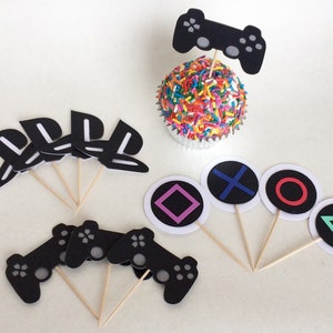 May include: A collection of black and white video game-themed cupcake toppers. The toppers include controller shapes, the PlayStation logo, and various colored geometric symbols. A cupcake with rainbow sprinkles is also featured.