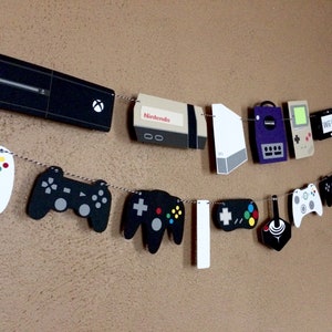 May include: A string of gaming console and controller cutouts, including a white Xbox, a black Nintendo, a white Wii, a purple GameCube, a green Game Boy, a white Wii, a black PS2, and several black and white controllers.