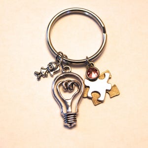 May include: Silver key chain with a light bulb charm, a puzzle piece charm, a small girl charm, and a pink rhinestone.