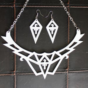 May include: A white geometric necklace and matching earrings set. The necklace features a chain and a large, openwork design. The earrings are diamond-shaped with a similar geometric pattern. The jewelry is displayed on a dark, textured surface.