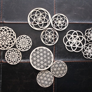 Puede incluir: Un conjunto de 10 posavasos de madera blanca geométricos mandala con diseños intrincados. Los posavasos están dispuestos en un patrón circular sobre una superficie negra.