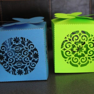 May include: Two decorative boxes, one blue and one green, with intricate circular cut-out designs. Each box has a butterfly-shaped accent on the lid. The boxes are cube-shaped and appear to be made of paper or cardstock.