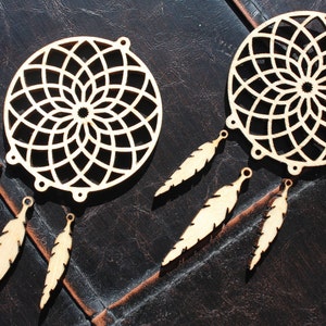 Op de afbeelding: Twee houten dromenvangers met ingewikkelde cirkelvormige ontwerpen en bevestigde veervormige hangers. De dromenvangers zijn gemaakt van lichtgekleurd hout en worden gepresenteerd op een donker, getextureerd oppervlak. De veren zijn ook van hout.